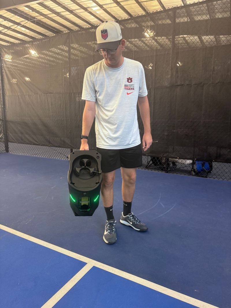 Coach Graham Richey holding the Tennibot Partner Lite by its handle, the feed wheel and loaded pickleballs visible from the front
