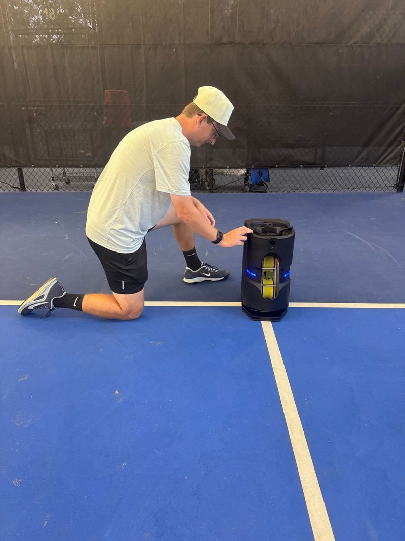 Coach Graham Richey kneeling on a covered pickleball court beside the Tennibot Partner Lite ball machine, pickleballs loaded in its window