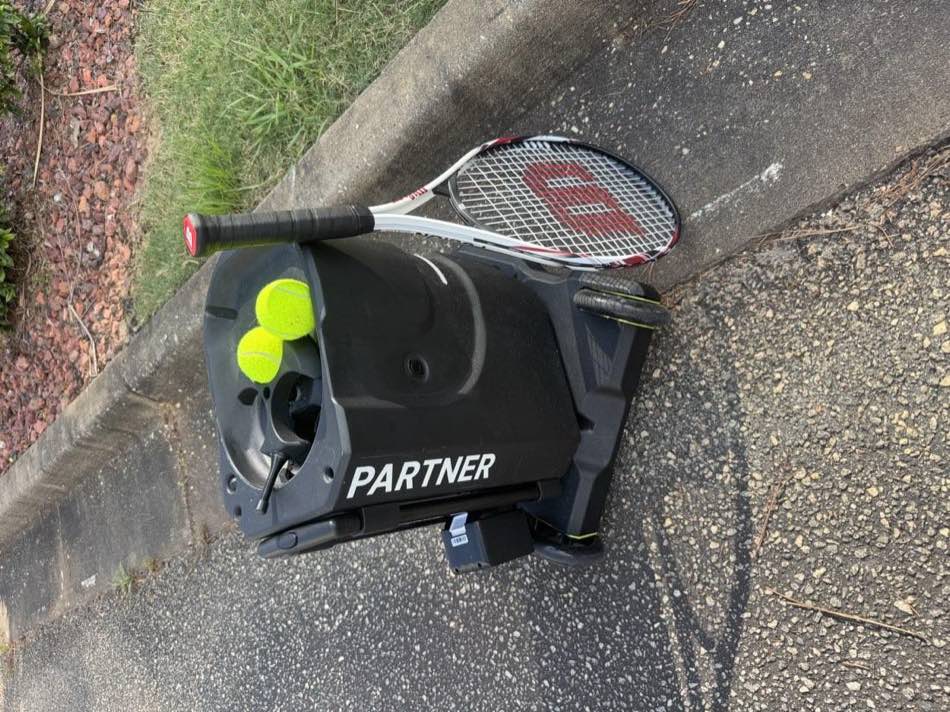 Tennibot Partner tennis ball machine loaded with tennis balls, a Wilson racquet leaning against it on a court path