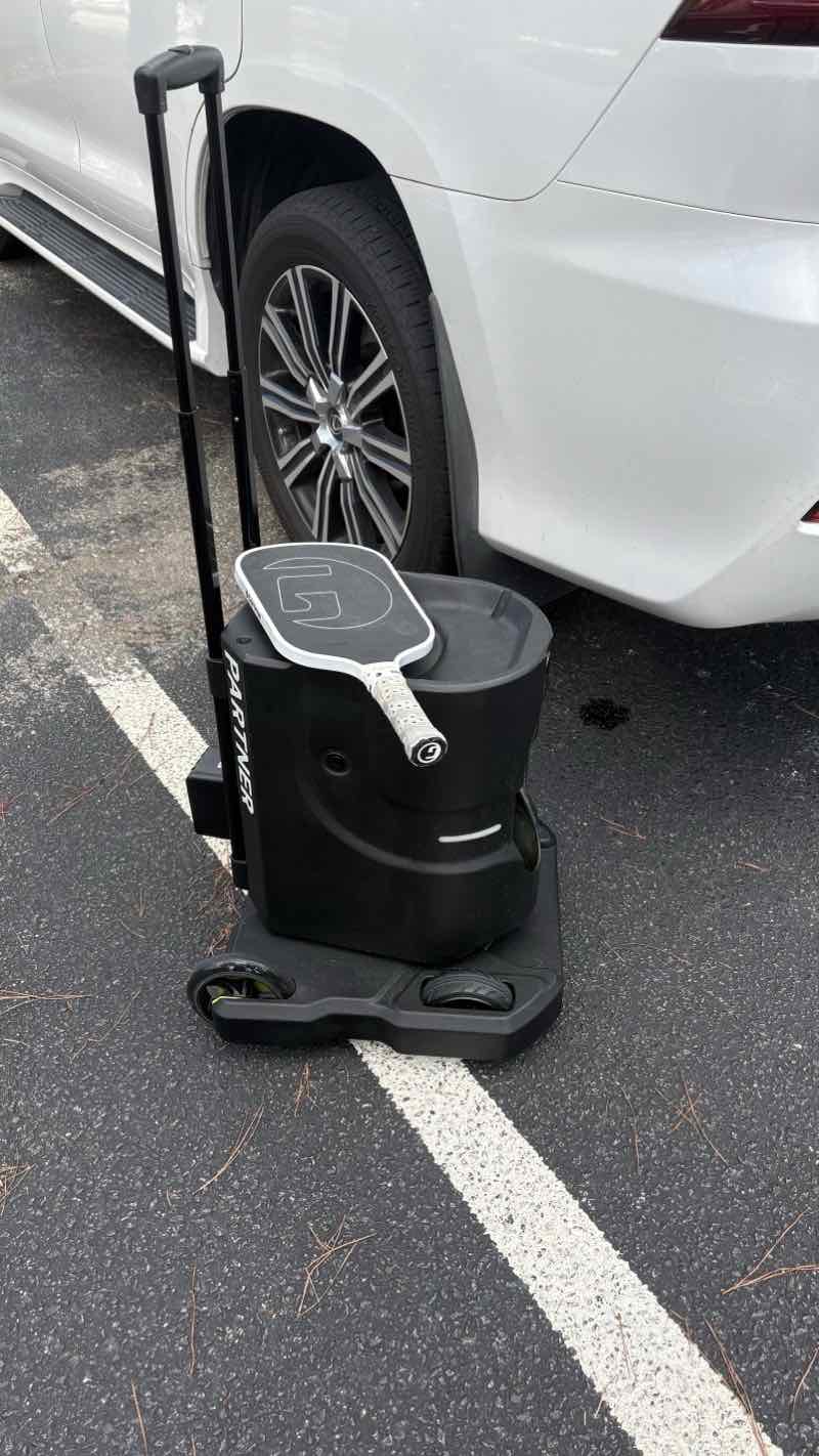 Tennibot Partner pickleball ball machine on its wheeled cart in a parking lot, a pickleball paddle resting on top